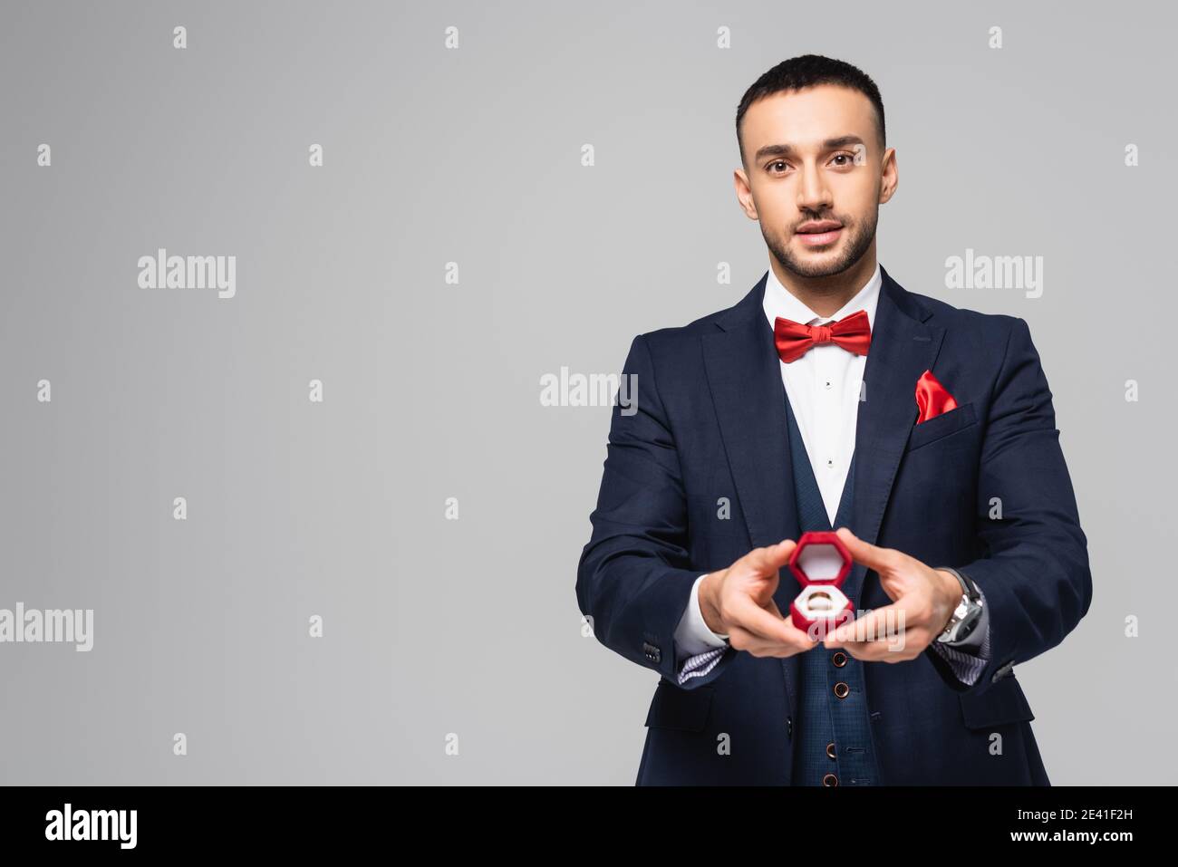 young hispanic man in elegant suit holding red jewelry box with wedding ring isolated on grey Stock Photo