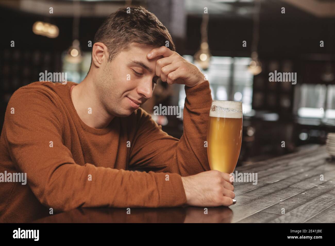 Young drunk man sleeping at the pub counter, smiling joyfully, holding full glass of delicious beer, copy space. Attractive tired man relaxing, drinki Stock Photo