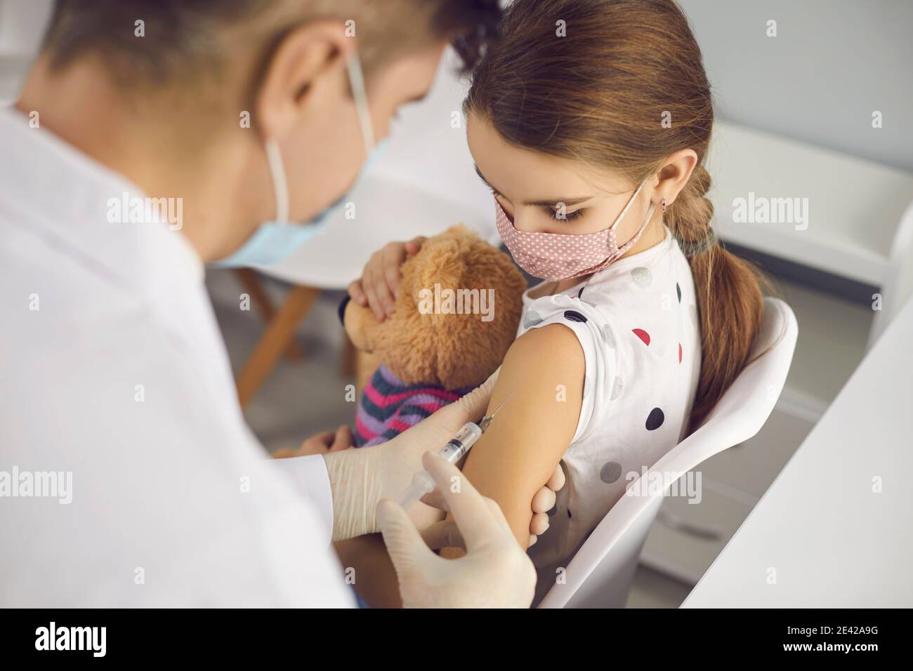 Little girl in protective medical face mask sitting with toy and getting vaccination Stock Photo