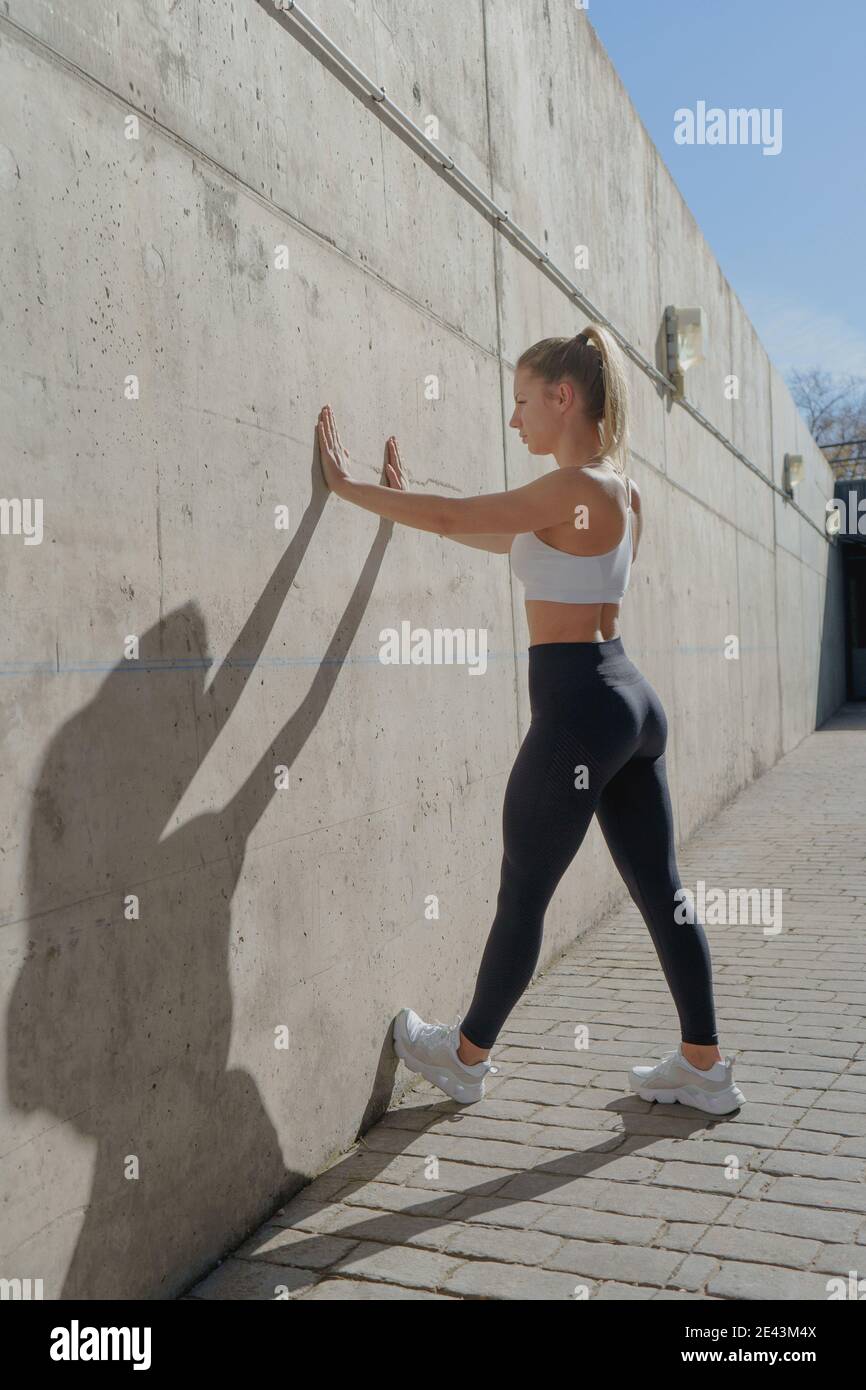 Side view of young fit Woman in sportswear doing standing push ups against concrete wall on street Stock Photo