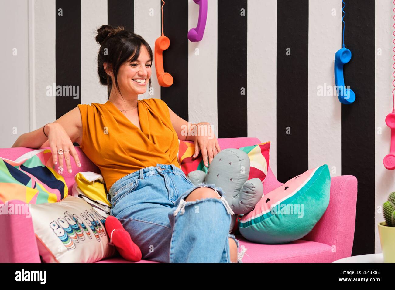 Young female in trendy ripped jeans laughing happily while resting on pink couch with colorful cushions in stylish room with retro telephone receivers Stock Photo