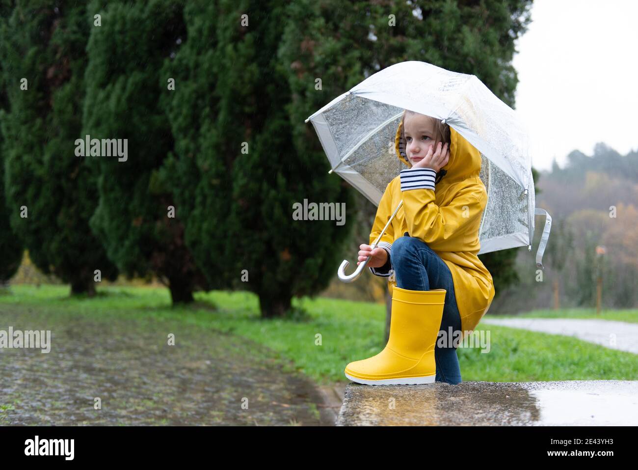 Tranquil kid in yellow raincoat and rubber boots standing under umbrella in  wet park on rainy day while leaning on hand and looking away Stock Photo -  Alamy