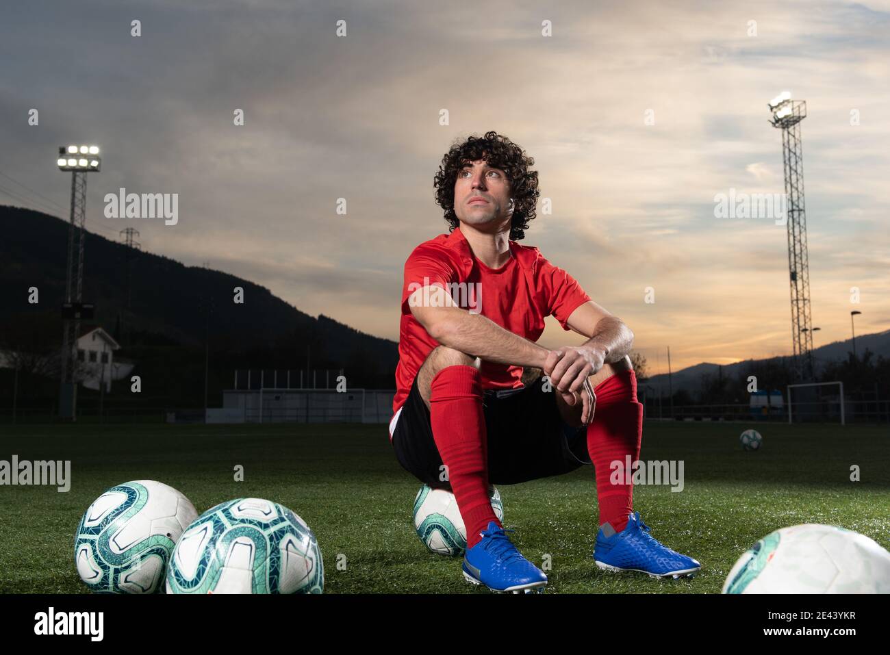 Low angle full body of serious determined sportsman in red soccer uniform sitting on field with football balls and looking away thoughtfully while res Stock Photo