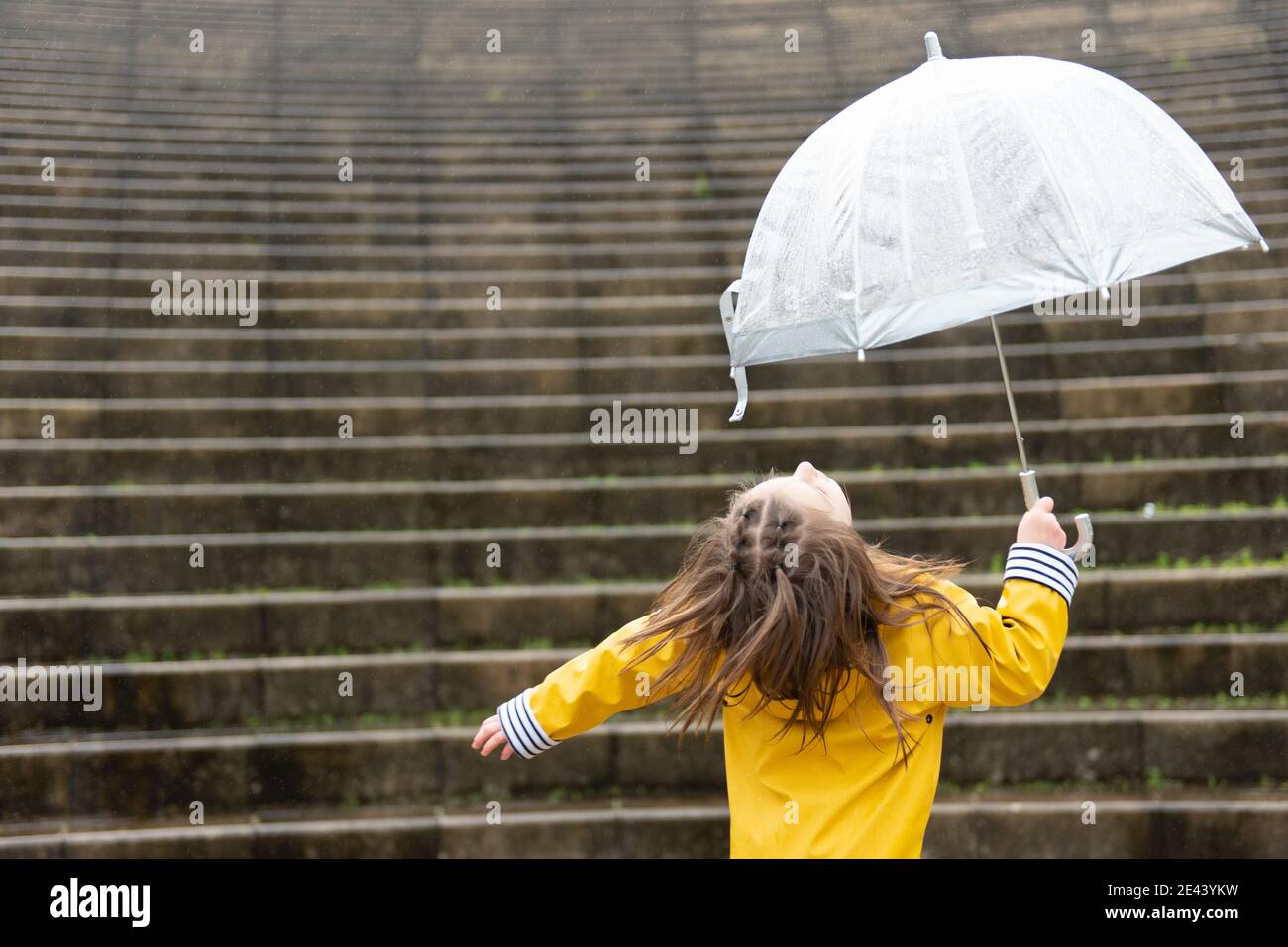 Back view of playful kid in yellow raincoat and with wet umbrella