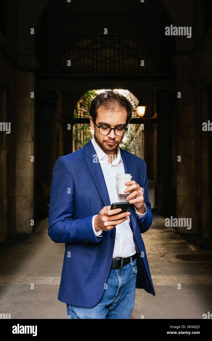 Serious businessman in smart casual outfit standing in city with cup of coffee to go and reading messages on smartphone Stock Photo
