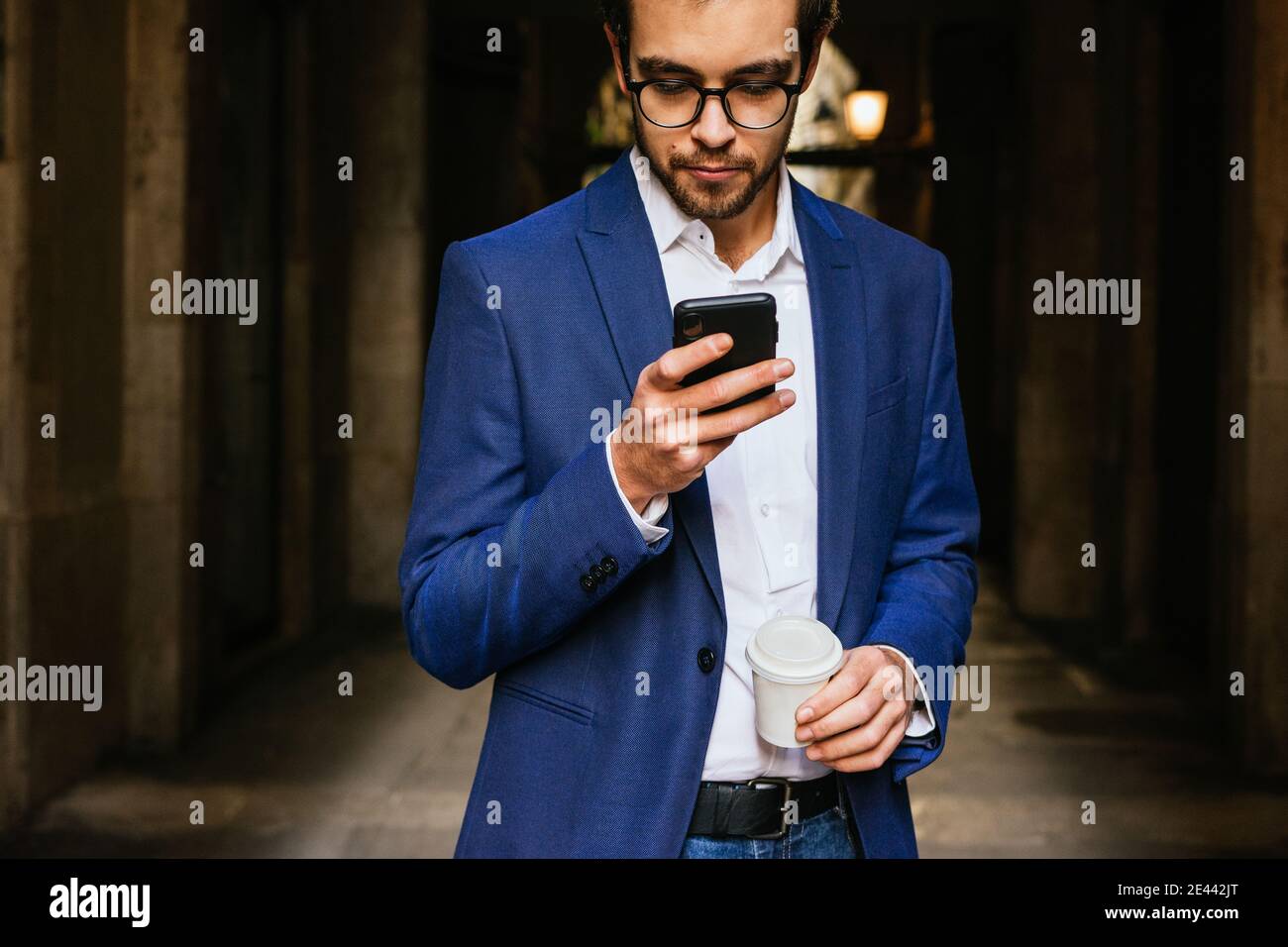 Serious businessman in smart casual outfit standing in city with cup of coffee to go and reading messages on smartphone Stock Photo