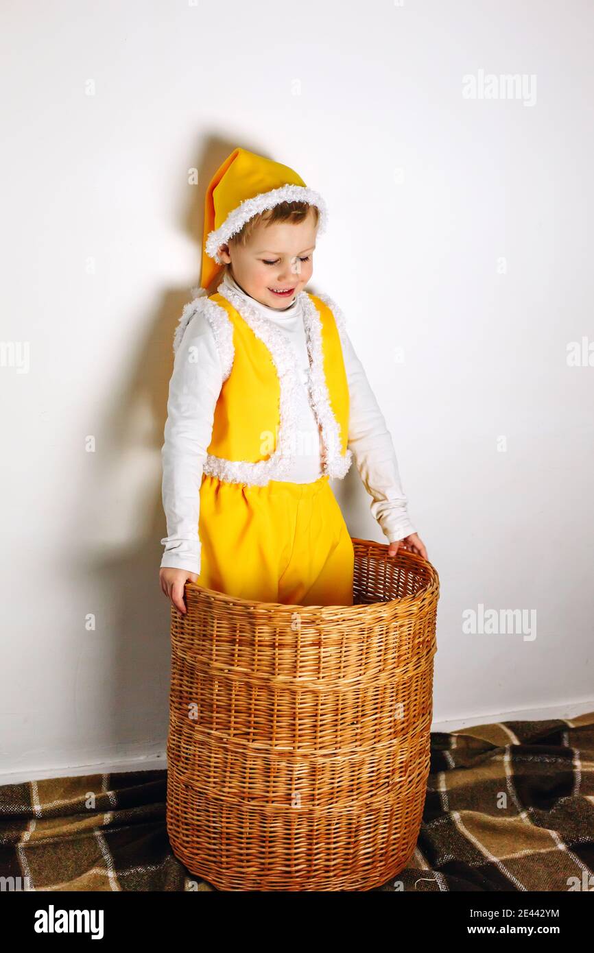 Cute smiling boy in yellow gnome elf outfit with funn hat, vest and cap stand inside straw wicker basket, family home fun Stock Photo