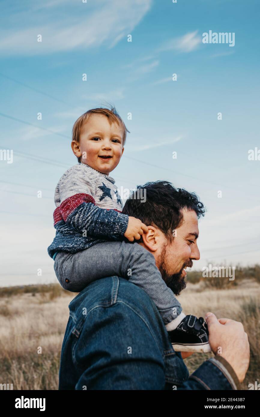 Side view of glad little boy riding on shoulders of smiling father during weekend in countryside Stock Photo