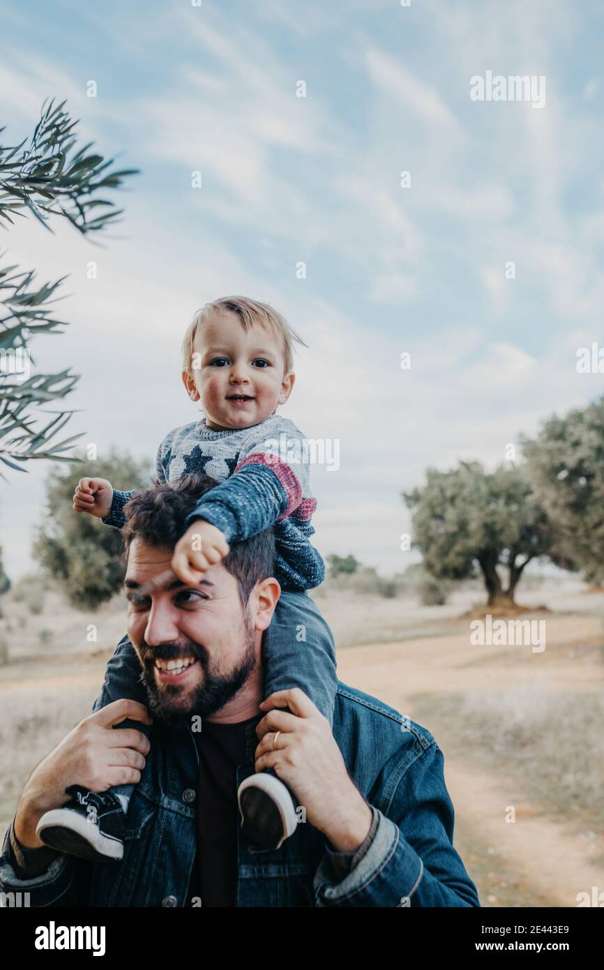 Side view of glad little boy riding on shoulders of smiling father during weekend in countryside Stock Photo
