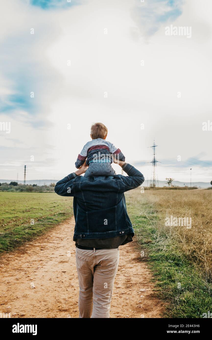 Back view of glad little boy riding on shoulders of smiling father during weekend in countryside Stock Photo