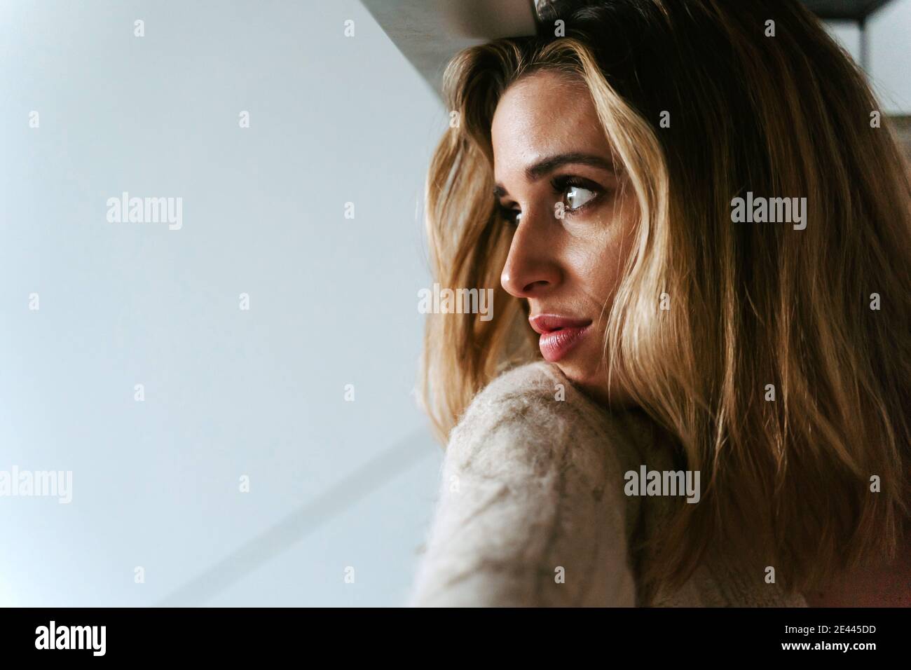 Charming thoughtful female with long hair and in cozy sweater looking out of window at home Stock Photo