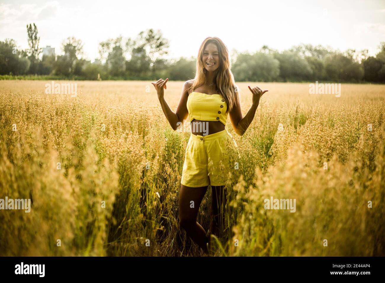 Cheerful young female in yellow summer wear showing shaka sign while standing in field in Salburua park and looking at camera Stock Photo