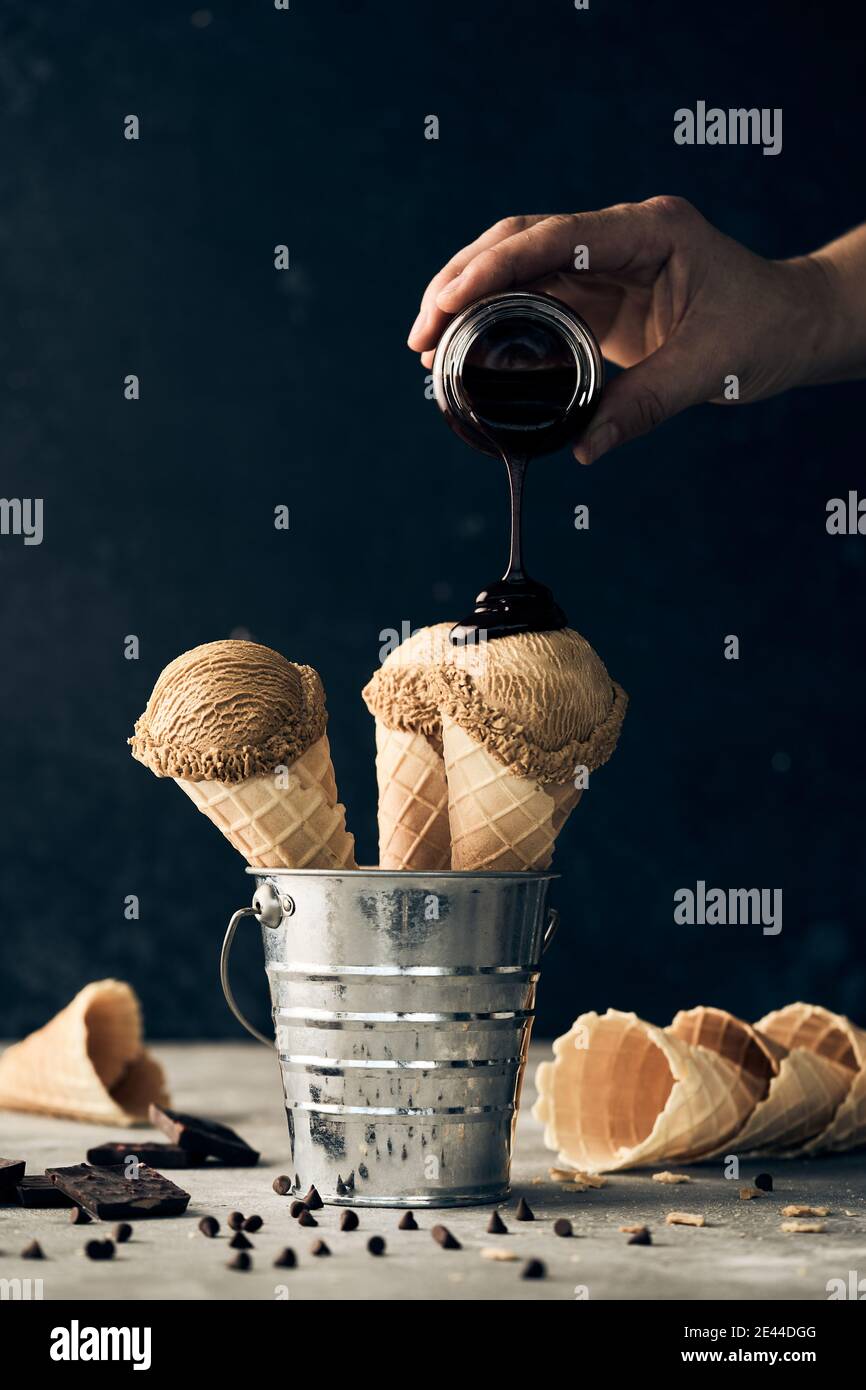 Crop anonymous person adding chocolate topping on scoops of ice cream in waffle cones placed in bucket on table on dark background Stock Photo