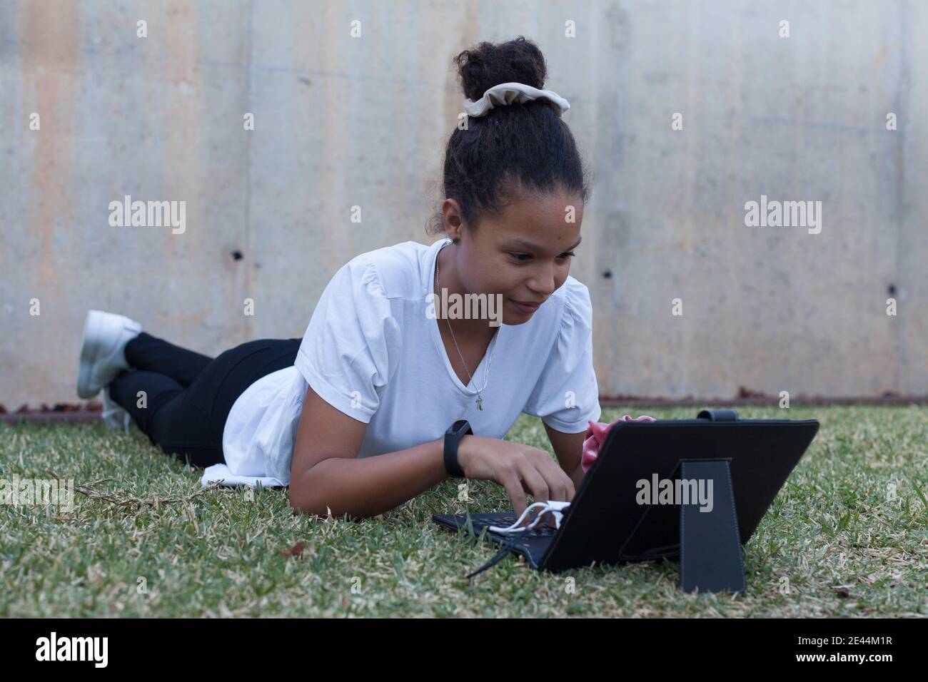 Full body of focused young African American female student in white t shirt lying on grassy lawn and typing on keyboard of modern tablet during remote Stock Photo