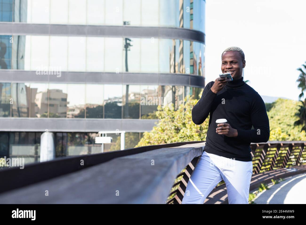 Confident young African American guy in stylish outfit using voice assistant while talking on mobile phone resting on building terrace with cup of tak Stock Photo