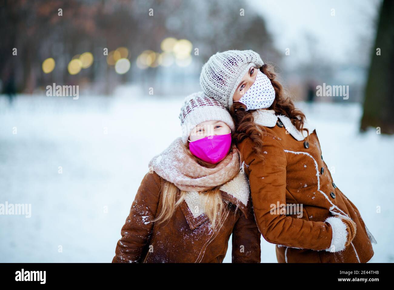 Portrait of smiling stylish mother and daughter in a knitted hats and sheepskin coats with medical mask in a knitted hat and sheepskin coat outside in Stock Photo