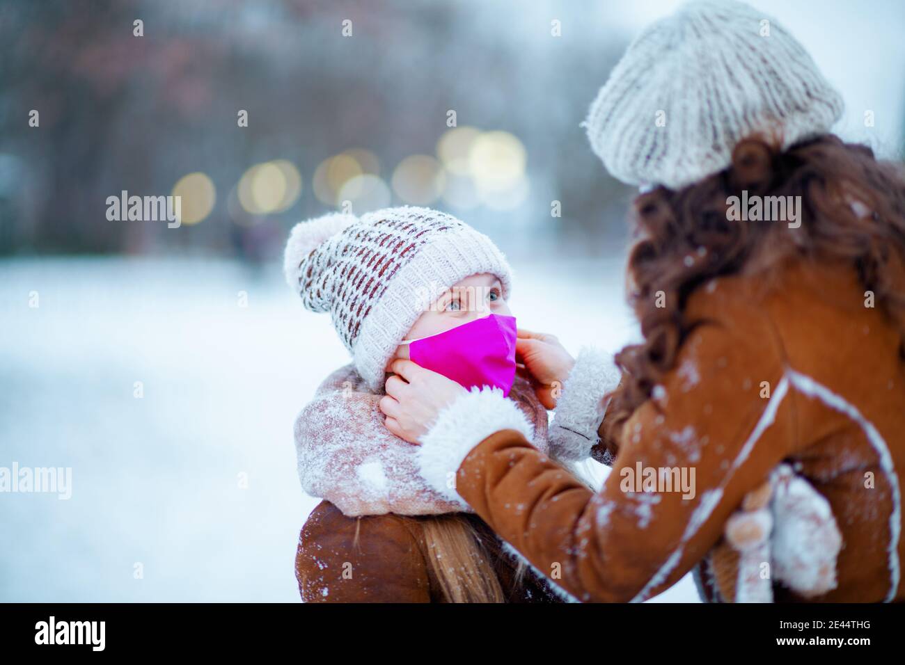 stylish mother and daughter in a knitted hats and sheepskin coats with medical mask in a knitted hat and sheepskin coat wearing medical mask outdoors Stock Photo