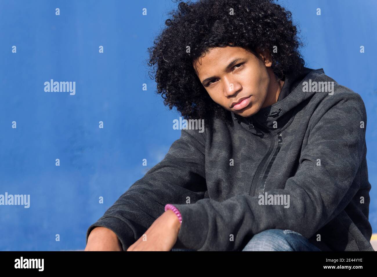 Thoughtful curly haired black male teenager in informal wear looking at camera while sitting against blue wall on street Stock Photo