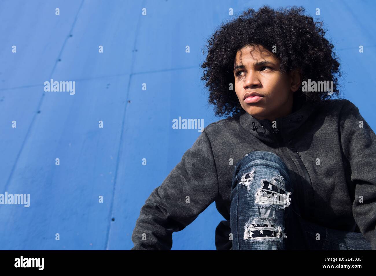 From below of thoughtful curly haired black male teenager in informal wear looking away while sitting against blue wall on street Stock Photo
