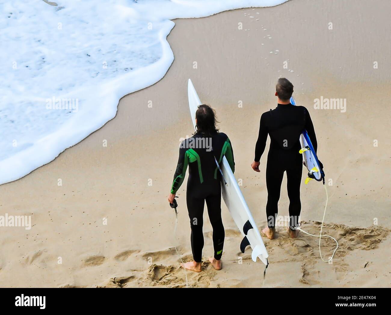 Two male surfers in black wetsuits holding their surfboards on a sandy