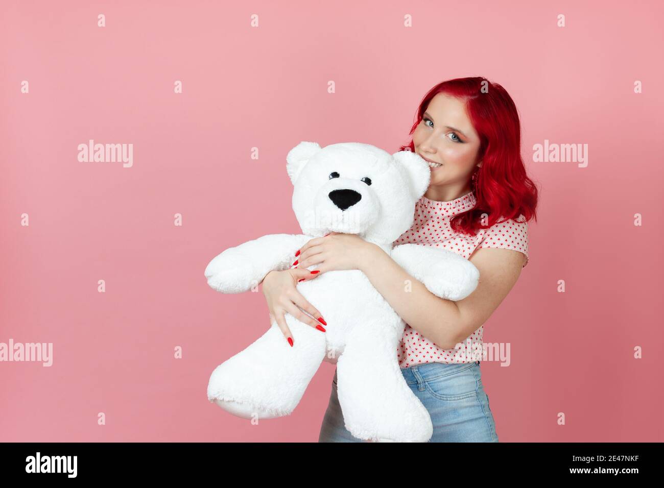 Close-up naughty, playful woman with red hair bites the ear of a large white teddy bear isolated on a pink background Stock Photo