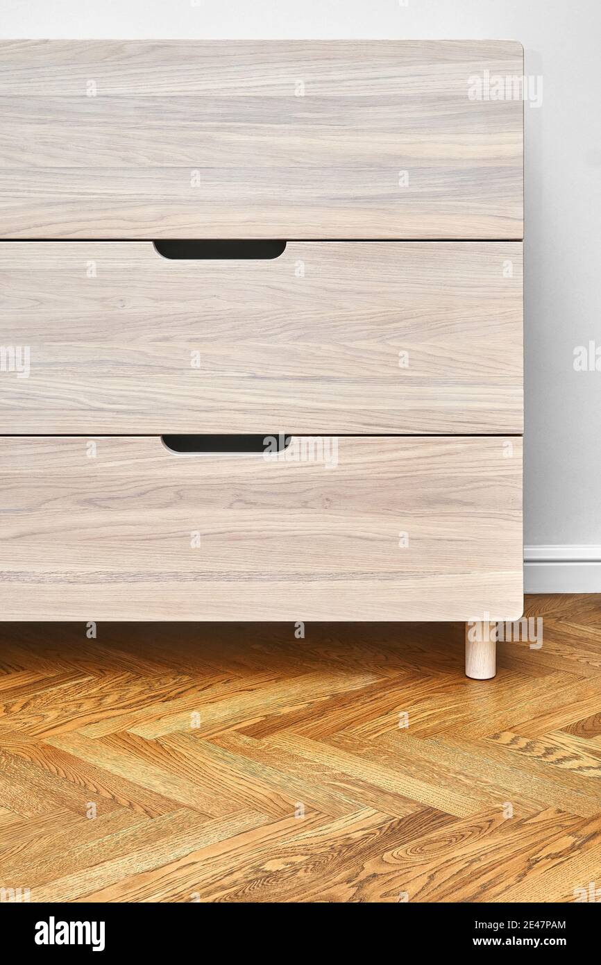 Stylish chest of drawers made with bleached solid oak timber with finger pull sliding stands on parquet floor near white wall Stock Photo