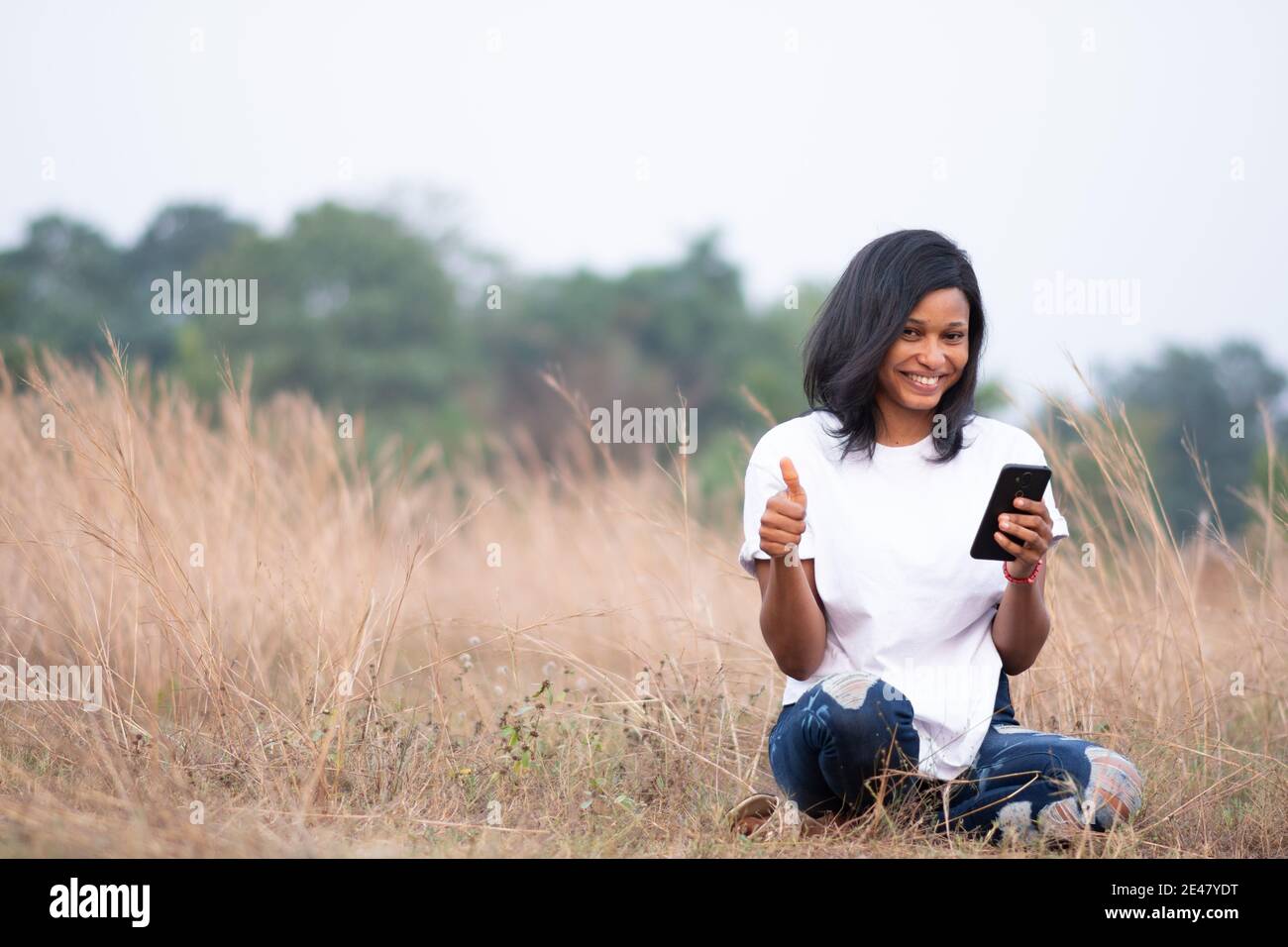 beautiful light skin african woman is smiling while using her phone and gives thumbs up towards the camera Stock Photo
