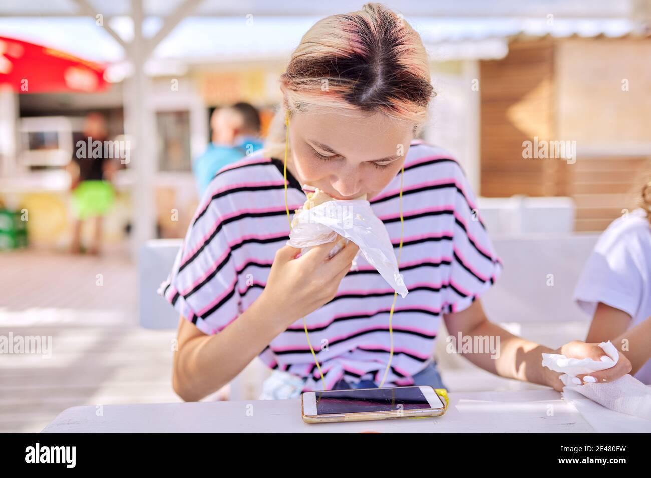Teen girl eating a sandwich, burger, looking at the smartphone screen in headphones Stock Photo