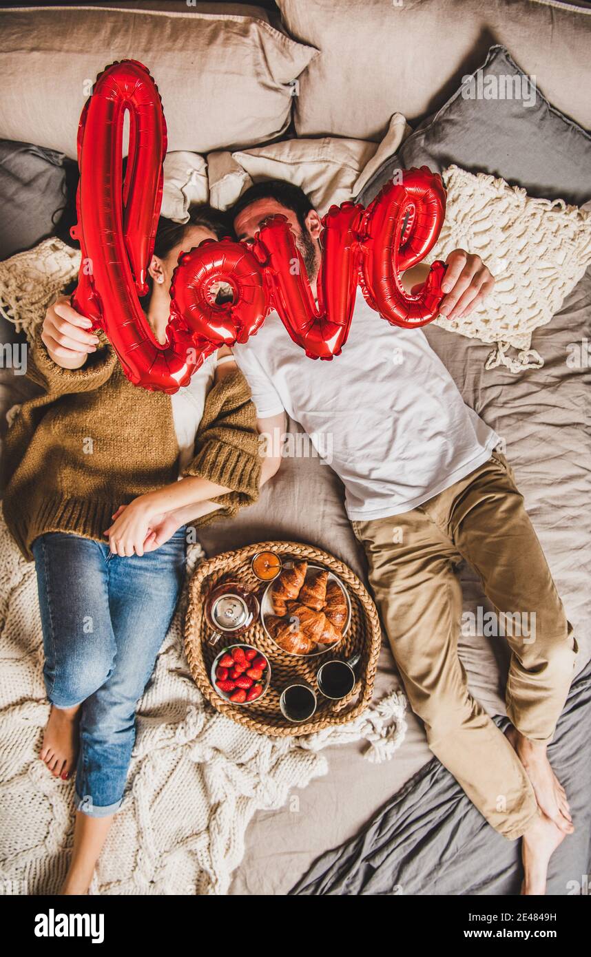 Young happy couple lying on pastel colored blankets barefoot in bed, having breakfast with fresh croissants, strawberries and tea, holding hands and sign love, top view. Saint Valentines day concept Stock Photo