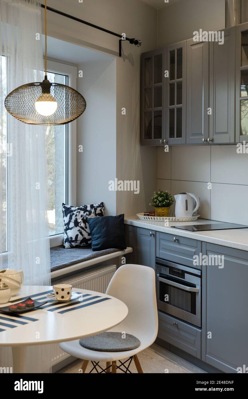 Interior of the newly equipped kitchen part in the studio apartment. Round table with lamp, chair by the window and wide windowsill with cushions. Kit Stock Photo
