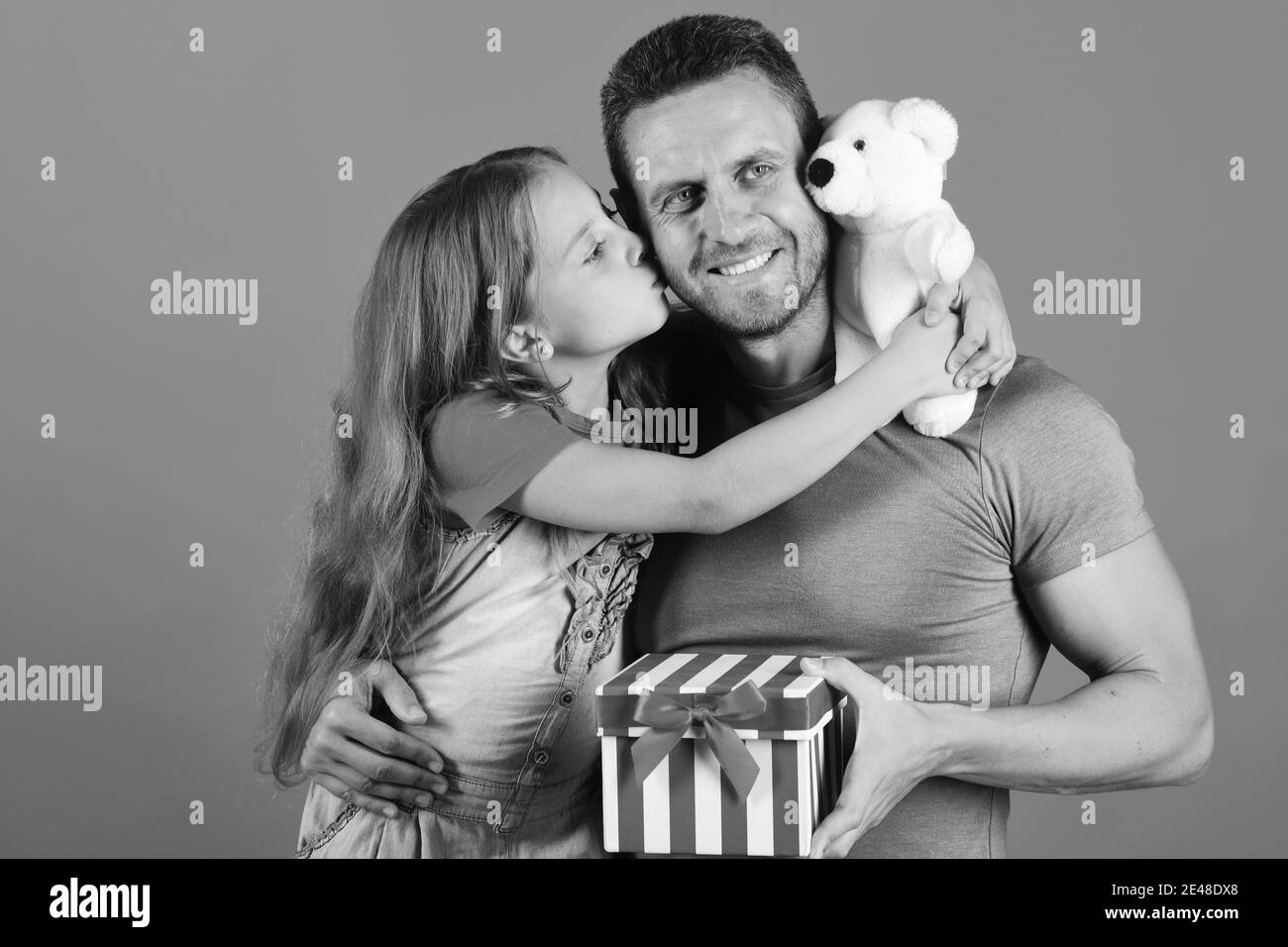 Girl and man with happy faces on red background. Kid kisses her daddy. Schoolgirl and dad hold present box and white teddy bear. Childhood, surprise gift and family concept. Daughter and father hug. Stock Photo