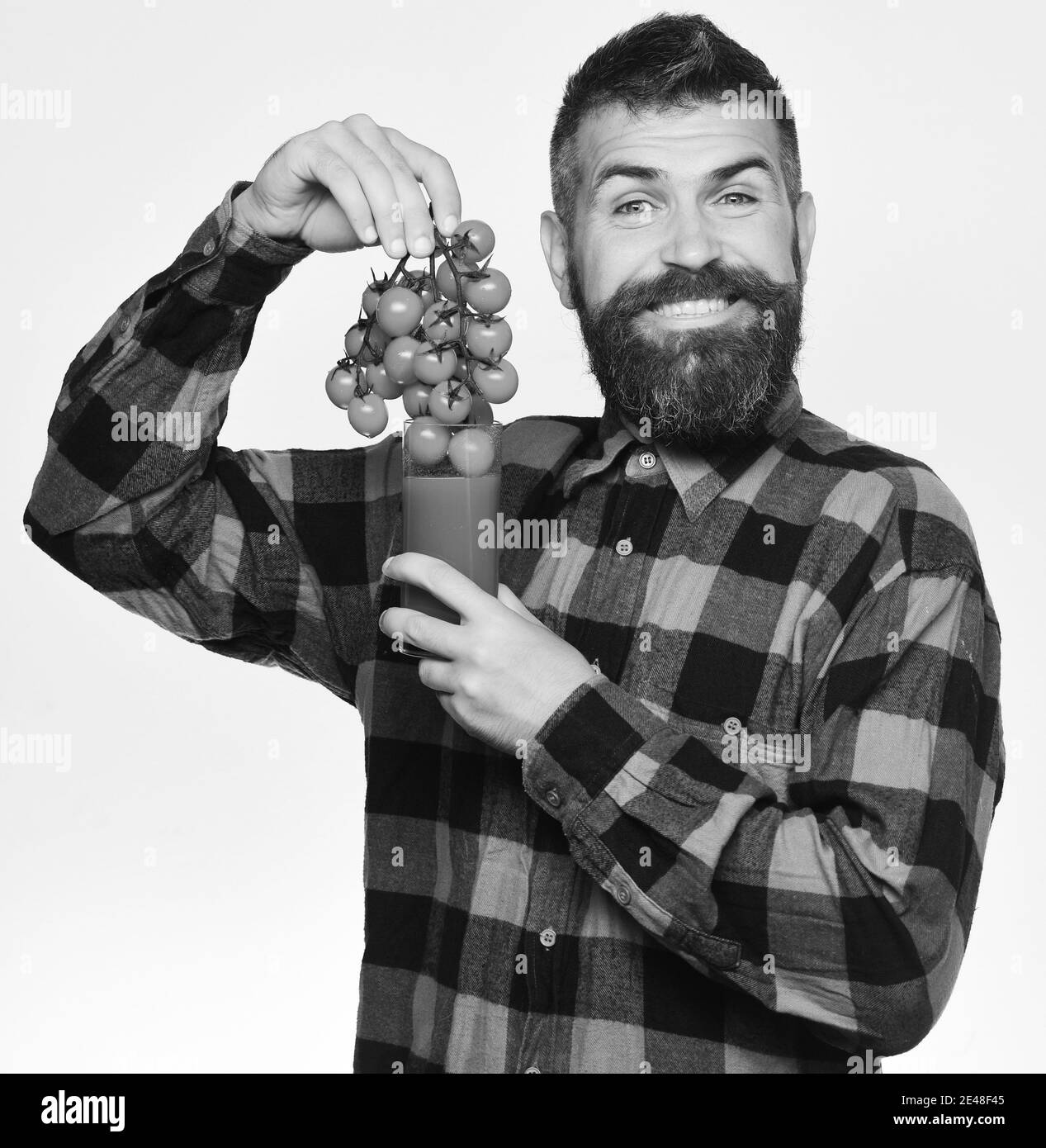 Farming and autumn concept. Guy with harvest. Farmer with smiling face offers fresh juice made of cherry tomatoes. Man with beard puts berries into glass of tomato juice isolated on white background. Stock Photo