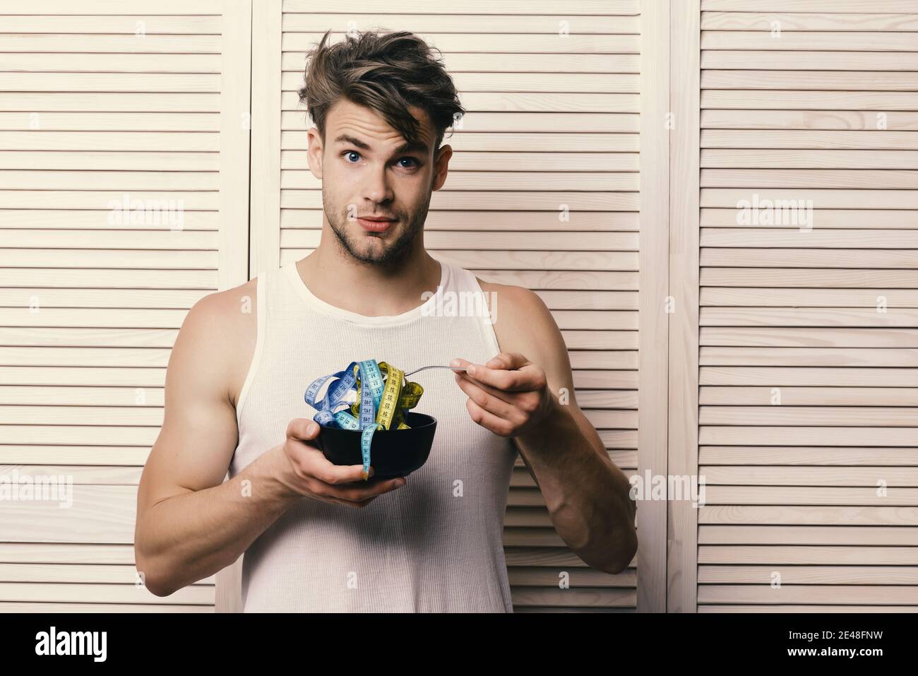 Diet and health concept. Athlete with messy hair and unshaved face. Guy in  sleeveless shirt eating measuring tape. Man with surprised face expression  on background of beige jalousie Stock Photo - Alamy