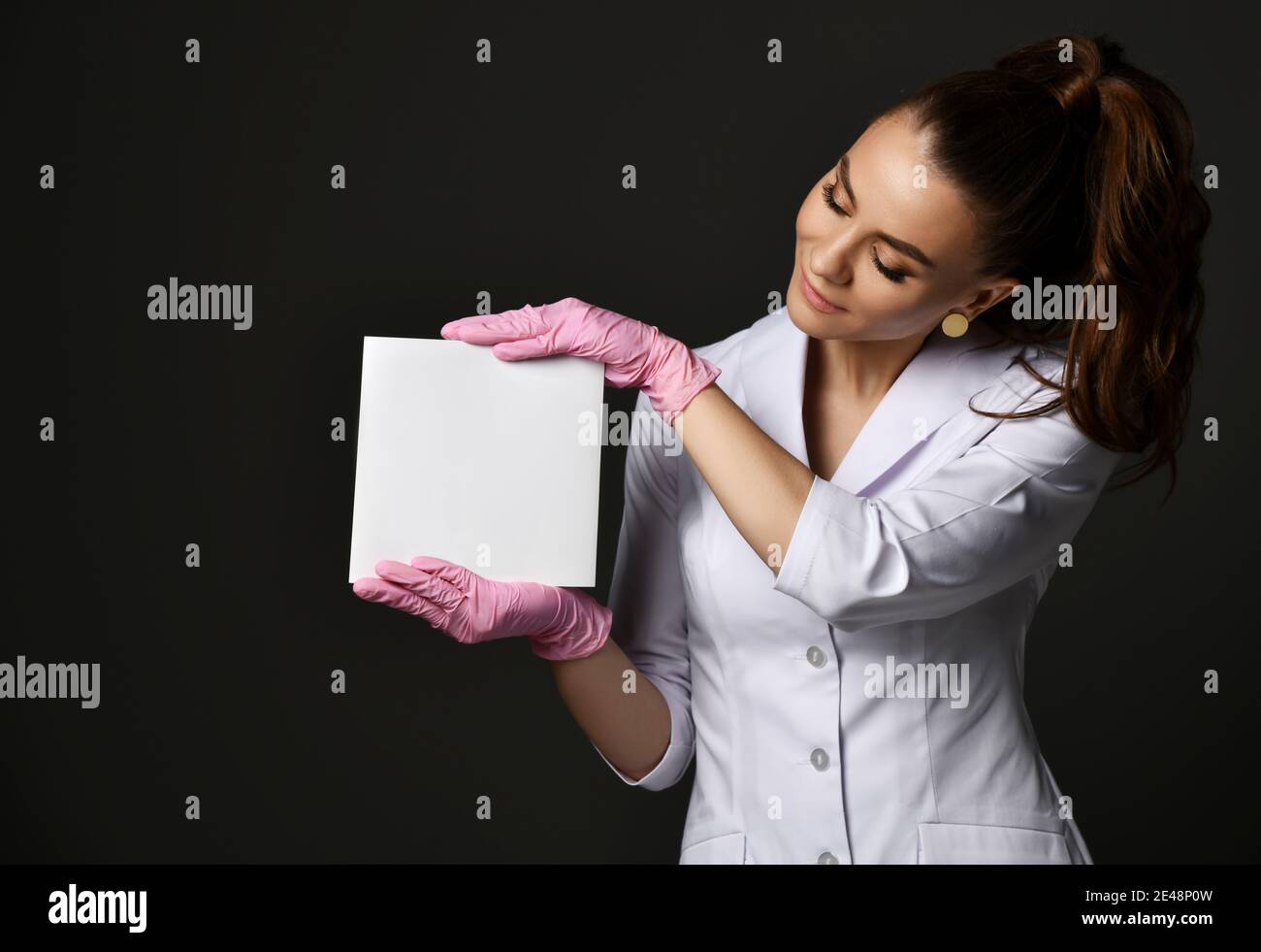 Young smiling pretty woman doctor therapist in white uniform and protective latex gloves holding blank paper sheet Stock Photo