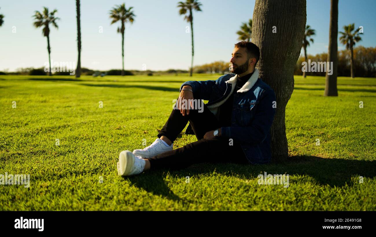 Young guy sitting under tree on green field outdoors Stock Photo