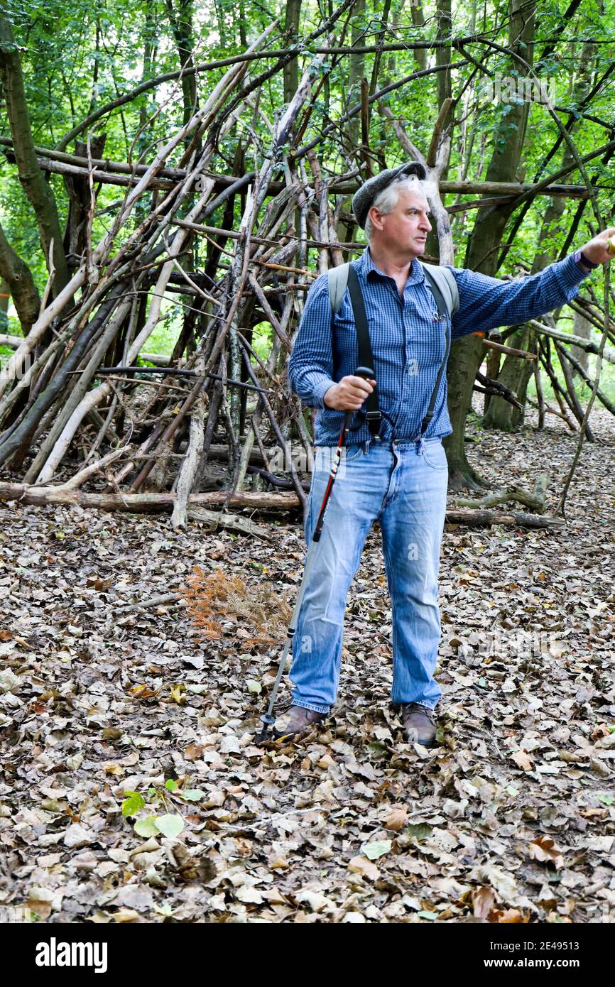 White haired, caucasian male wearing suspenders, beret and blue jeans i woods in front of a fort made out of branches Stock Photo