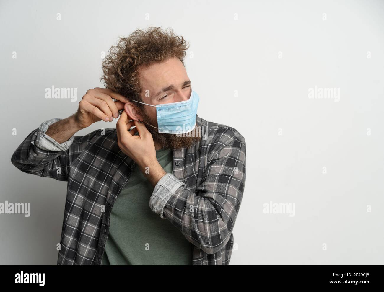 Putting on or taking off protective sterile medical mask on his face to protect coronavirus, with curly hair, young man wearing plaid shirt and olive Stock Photo