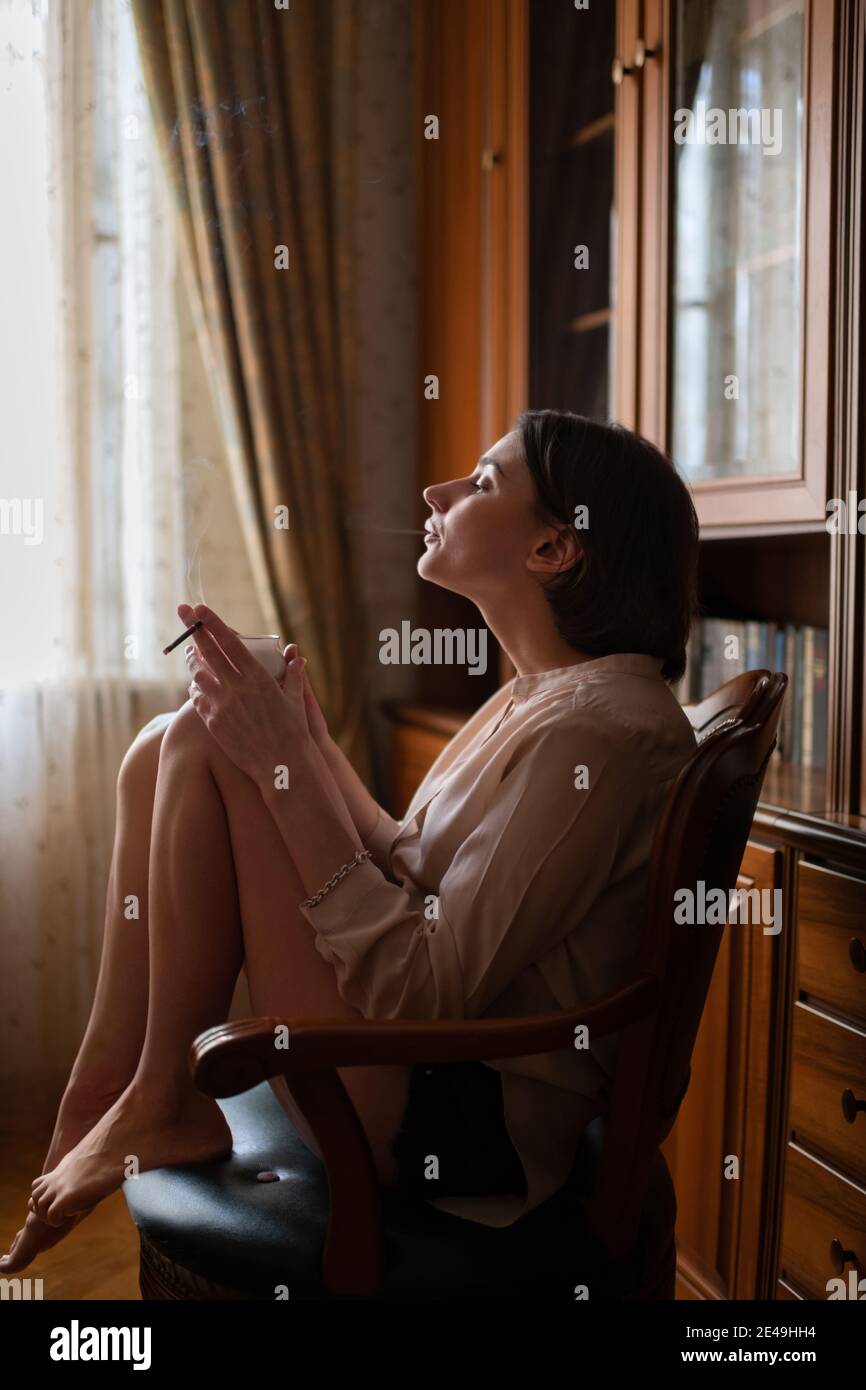 Side view of woman with hot drink exhaling cigarette smoke while resting at home Stock Photo