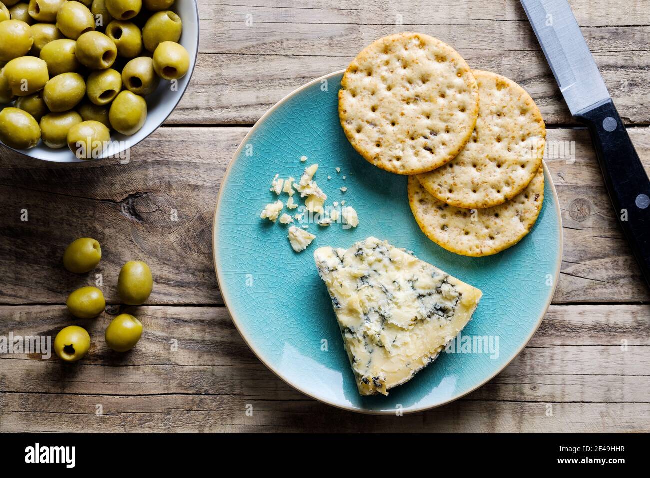 A slice of mature Stilton cheese on a plate with round wholewheat crackers with some green olives in a small bowl to the side Stock Photo
