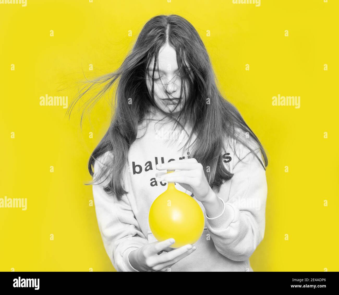 Black and white portrait of young teenage girl with flying hair with yellow balloon blowing air into her face against background in studio. Color Conc Stock Photo