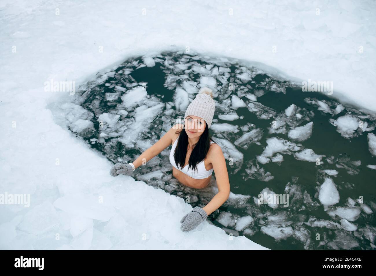 Winter swimming. Woman in frozen lake ice hole. Swimmers wellness in icy water. How to swim in cold water. Beautiful young female in zen meditation. Stock Photo