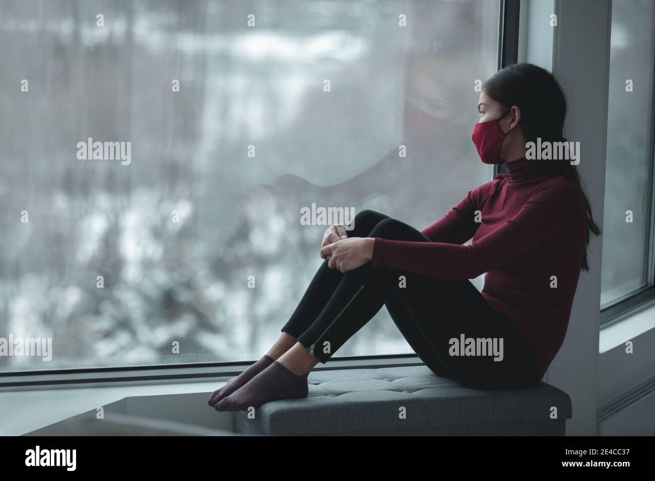 Winter depression because of coronavirus confinement. Sad Asian woman alone during city lockdown wearing face mask indoors at home covid. Anxiety Stock Photo