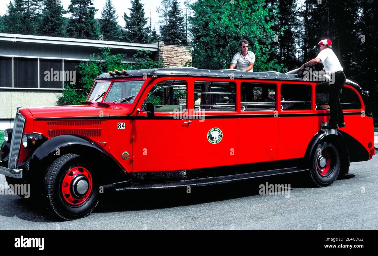 Two men on this long 1930s red touring bus open its roll-back canvas rooftop to provide panoramic views of awesome mountain peaks for tourists taking scenic excursions in Glacier National Park in northwest Montana, USA. The 17-passenger vehicle is among the park’s fleet of 33 identical buses that are known as Red Jammers because their drivers noisily 'jammed' the gears when double-shifting the manual transmissions on the steep mountainous roads. All of those historic White Motor Company No. 706 models have been restored to their original appearance and retrofitted with automatic transmissions. Stock Photo