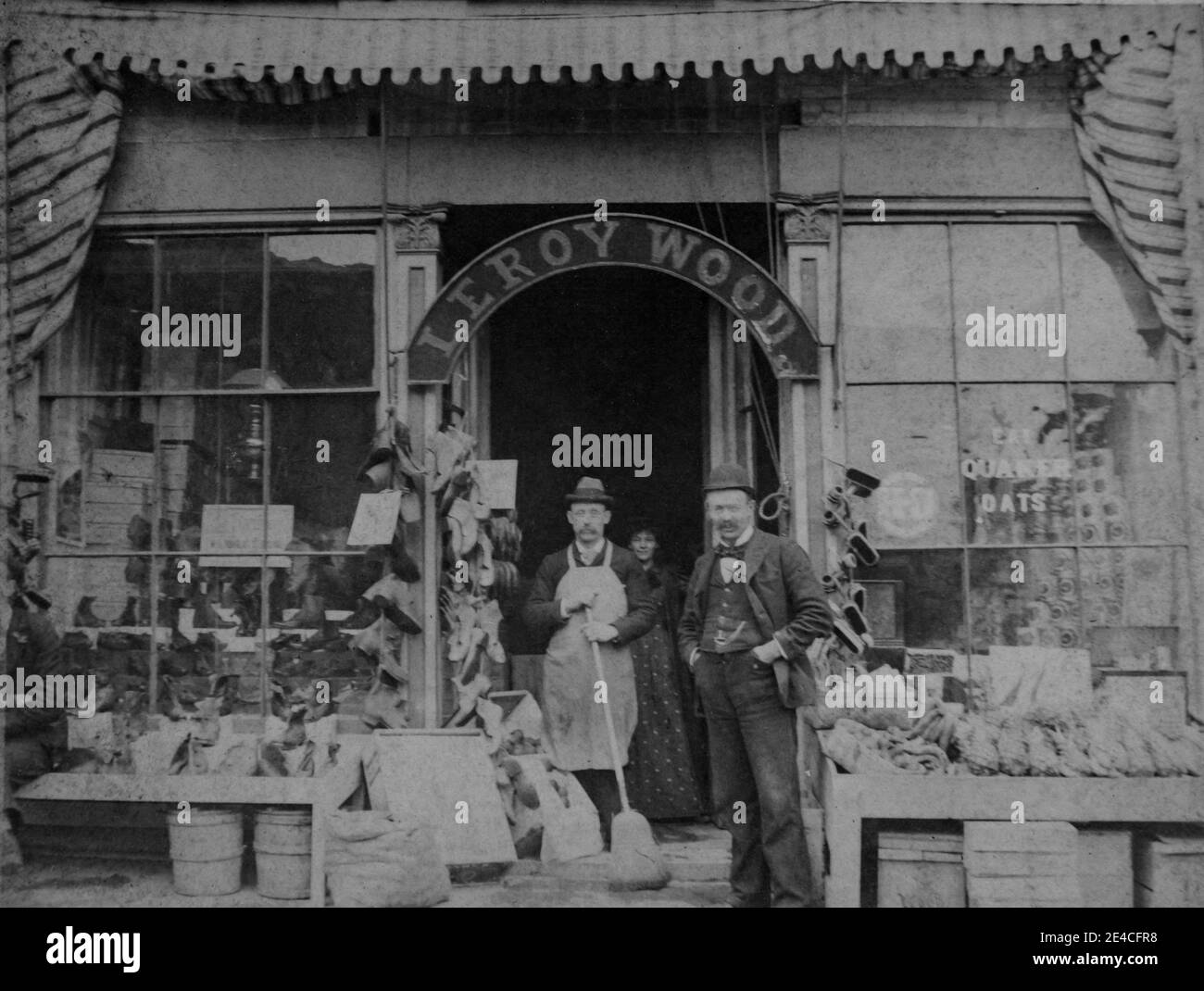 American archive monochrome portrait  of a grocer shop or general store front with an arched sign with name Leroy Wood and an advert in the window saying  Eat Quaker Oats and merchandise for sale on display. Two men are wearing bowler hats. A man is holding a broom, and a woman is standing behind the men. Taken in the late 19th century in NY, USA Stock Photo