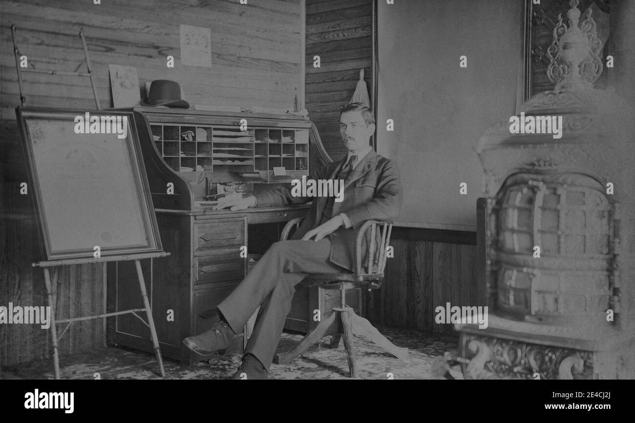 American archive monochrome portrait  of a man in a suit sitting at a roll top desk in an office with an easel and a large furnace, USA Stock Photo