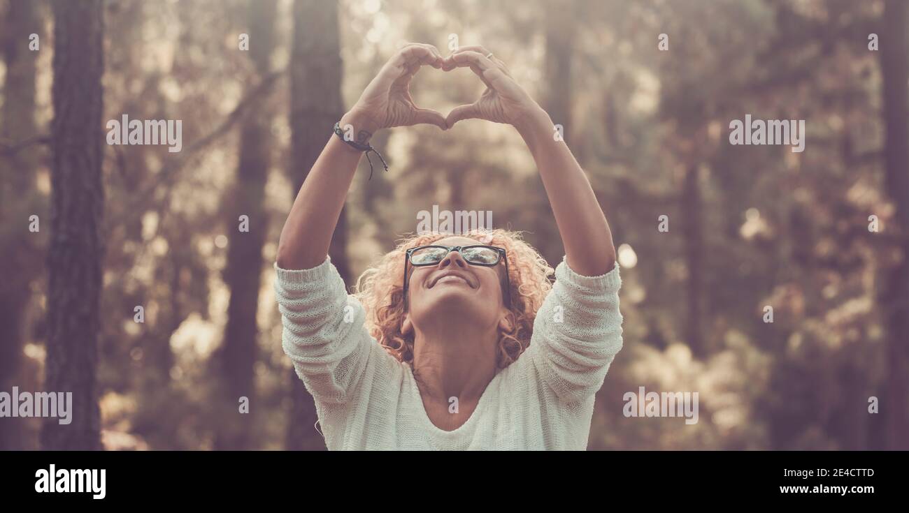 Cheerful woman enjoy nature and outdoors natural forest doing hearth sign with hands and smiling to trees - concept of save nature world and environment lifestyle people Stock Photo
