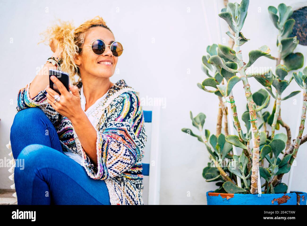 Cheerful beautiful woman portrait use cellular phone outdoor with plant and blue seats outdoor home - people enjoying life and technology concept Stock Photo