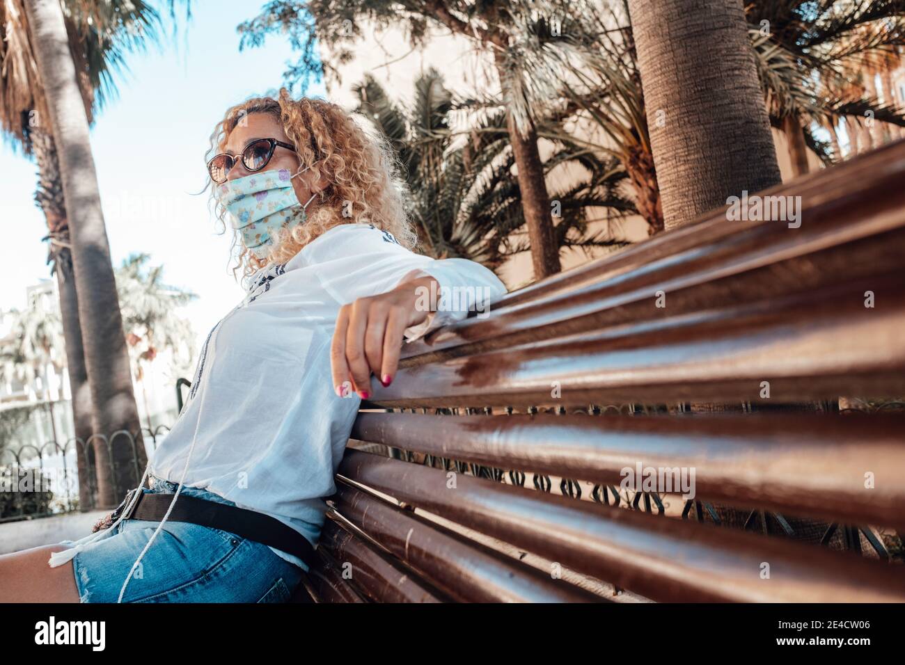 Woman sit down on a bench wearing medical face mask for coronavirus covid-19 protection outbreaks - concept of outdoor restrictions to fight pandemic contagion for health of people Stock Photo