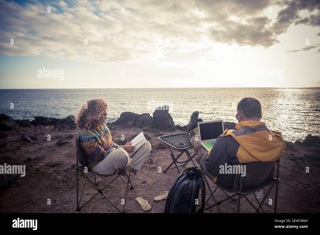 Adult couple sit down on the rock beach with computer and book enjoying the travel adventure vacation and smart working online concept - people and outdoors lifestyle Stock Photo