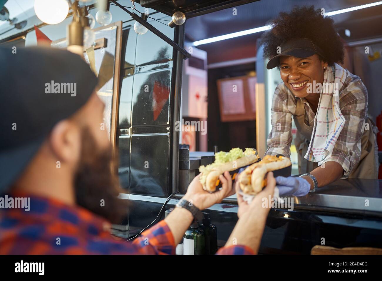 beautiful young afro-american female employee looking at camera, smiling, while giving sandwiches through a fast food window to a caucasian male custo Stock Photo